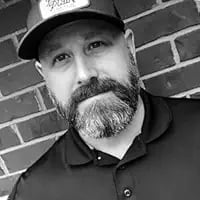 Black and white portrait of a bearded man wearing a baseball cap and polo shirt against a brick wall