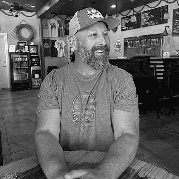 Bearded man wearing a cap and graphic t-shirt sitting in a shop interior in black and white